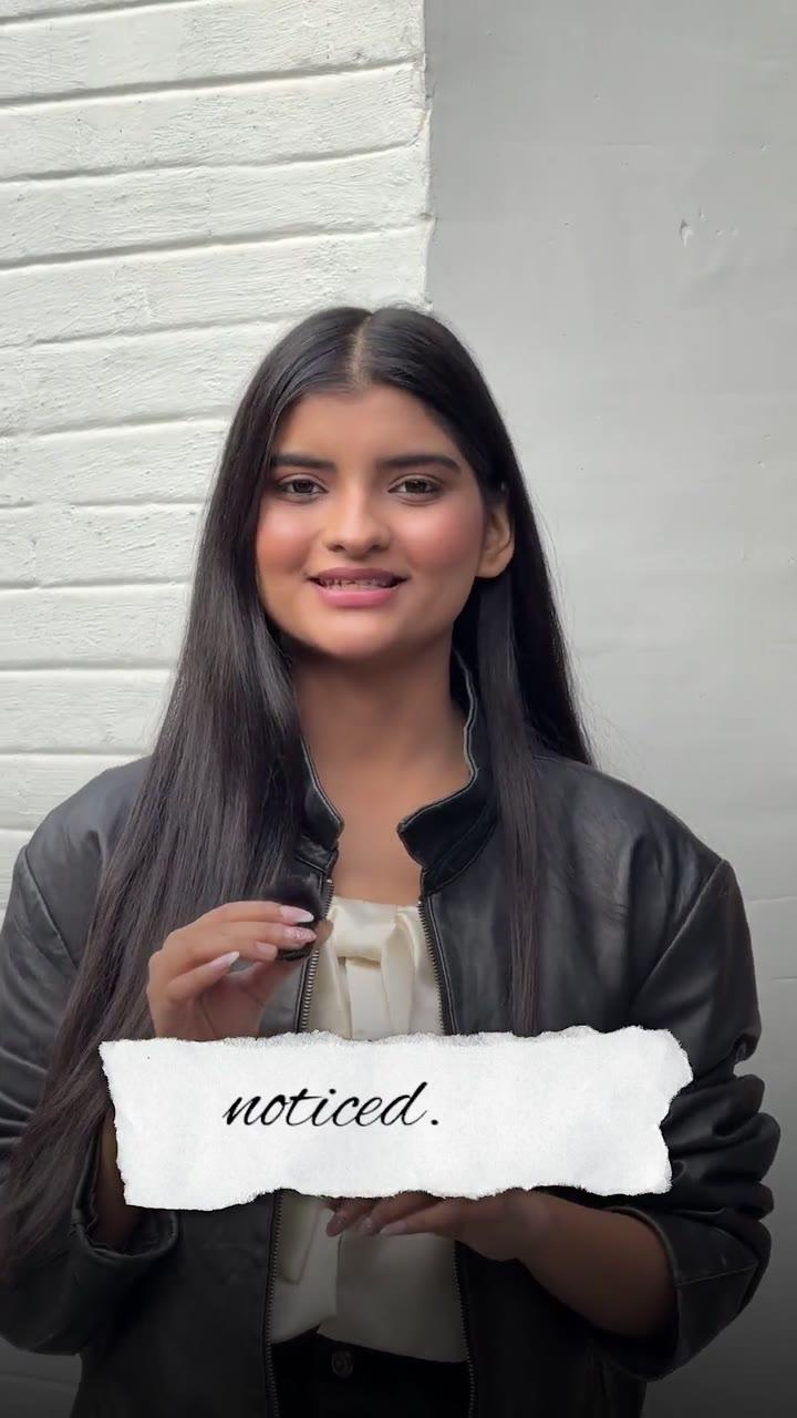 Frame from the CollabX UGC video: a woman in a black leather jacket and white top speaking to the camera outdoors against a white brick wall, holding a torn paper style caption label reading noticed.