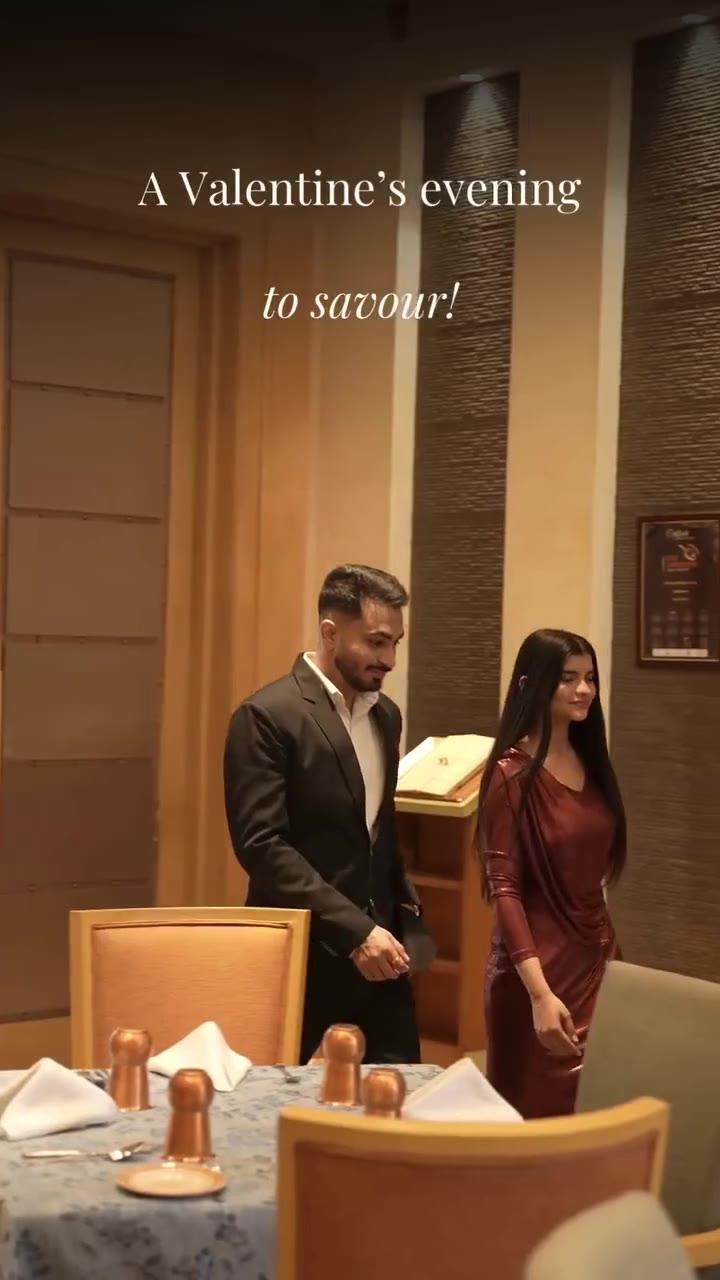 Frame from the DoubleTree by Hilton Agra campaign video: a man in a dark suit and a woman in a maroon dress standing at a set restaurant table, with the on screen line A Valentine's evening to savour.