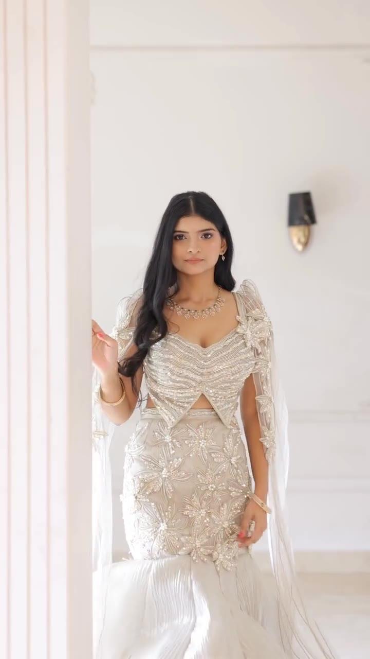 Frame from the VJ Jewellers campaign video: a model in an ivory and silver embroidered outfit with a necklace and earrings, standing beside a white column in a bright interior and looking at the camera.